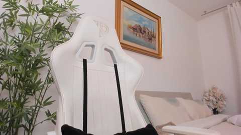 Video of a white gaming chair with black straps, placed in a minimalist bedroom with a framed painting of a coastal scene, bamboo plant, and beige bedding.