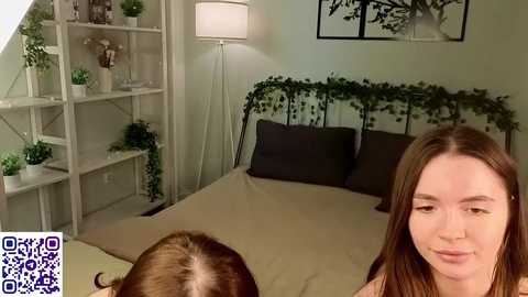 Video of a serene, green-themed bedroom with a woman and a dog on a beige bed; shelves with potted plants, a white lamp, and a black-framed tree wall art.