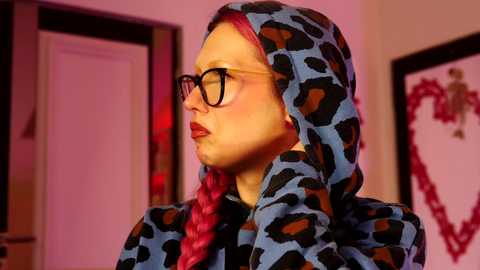 Video of a fair-skinned woman with long, bright pink hair, wearing glasses, a leopard-print hooded coat, and a braided hairdo, set against a warm, softly lit room with pink hues.