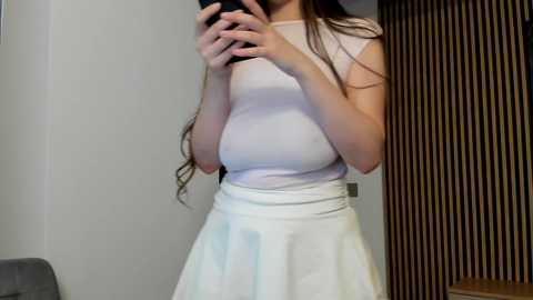 Video of a slim, fair-skinned woman with long, wavy brown hair, wearing a tight white top and high-waisted white skirt, standing indoors near a wooden door and gray couch.