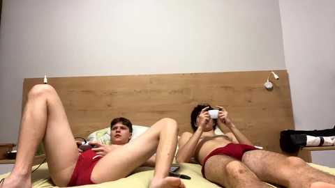 Video of two young men, one with fair skin and red briefs, lying on a bed, the other with dark skin and red briefs, holding a camera. Minimalistic bedroom background.