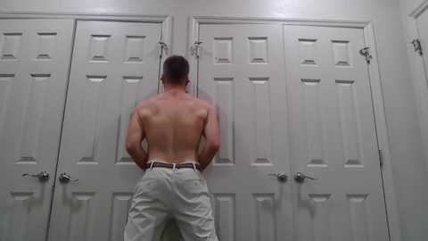 Video of a shirtless man with short brown hair, wearing white pants, standing with his back to white double doors, hands in pockets.