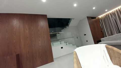 Video of a modern, minimalist bedroom with wooden wall panels, white furniture, and a glass staircase leading to an upper level.