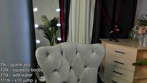 Video of a modern, light-grey, tufted armchair in a stylishly lit bedroom with a wooden dresser, potted plants, and red curtains.