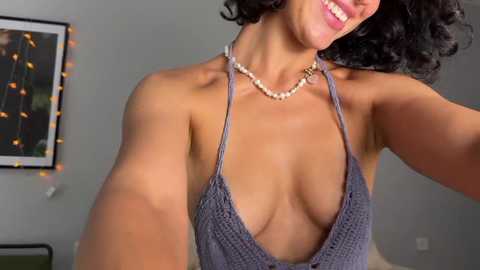 Media: Video of a smiling, light-skinned woman with curly black hair, wearing a low-cut, knitted gray halter top revealing cleavage, and a pearl necklace. Background shows a gray wall with a string of fairy lights and framed art.