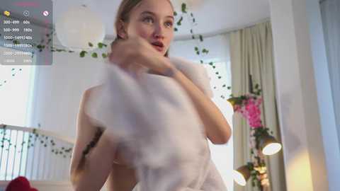 Video of a blonde woman with fair skin, wearing a white dress, hugging a fluffy white pillow, in a brightly lit, modern room with white walls, green plants, and a staircase.