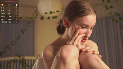 Video of a young, fair-skinned woman with brown hair, sitting nude, covering her face with her hands, in a dimly lit room with green vines, a radiator, and white curtains.