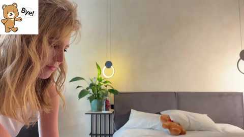 A video of a blonde woman with long hair and freckles, sitting on a bed with a teddy bear and potted plant, in a minimalist room with a gray headboard.