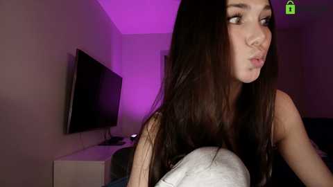 Video of a young Asian woman with long black hair, wearing a sleeveless top, making a kissy face. Background includes a TV, purple lighting, and minimalistic decor.
