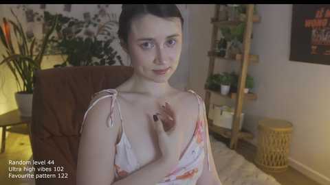 A video of a young, fair-skinned woman with dark hair, wearing a floral dress, seated in a dimly lit room. She holds her breast, revealing a nipple, while plants and a wooden ladder are in the background.