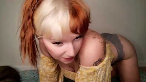 Video of a fair-skinned, red-haired woman with dyed blonde hair, wearing a yellow sweater and gray underwear, leaning forward with her face close to the camera, showing a tattoo on her shoulder.