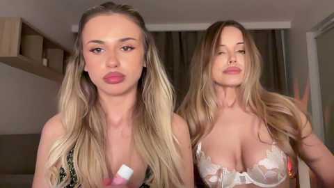 Media: Video of two young women with fair skin and long blonde hair, wearing lingerie. One has a pouty expression, the other smirks. Background features a minimalist bedroom with neutral tones.