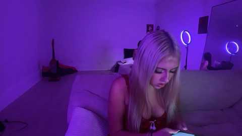Video of a blonde woman in a red bra, lying on a bed, engrossed in her phone, with purple lighting and a guitar in the background.