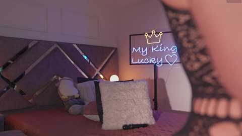 Video of a dimly lit bedroom featuring a bed with a plush pillow, a mirror, a neon sign reading \"My King, My Luxury,\" and a person wearing black lace lingerie, partially visible.