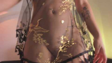 Video of a fair-skinned woman wearing sheer black lingerie adorned with gold floral embroidery, revealing her breasts and navel piercing. The background is softly lit with warm yellow tones.