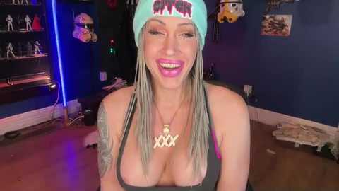 Video of a smiling, light-skinned woman with long blonde braids, wearing a blue beanie and a low-cut black top, revealing ample cleavage, in a cluttered, dimly lit room with blue walls.