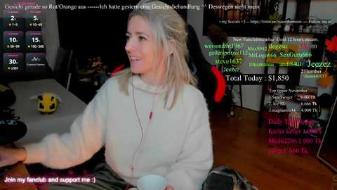 Video of a smiling, light-skinned woman with blonde hair in a gray hoodie, seated in a dimly lit room with a gaming setup and chat text overlay.