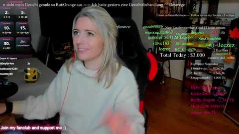 A video shows a blonde woman in a light blue sweater, sitting at a computer desk in a dimly lit room. Chat windows and gaming overlays are visible, suggesting she's livestreaming.