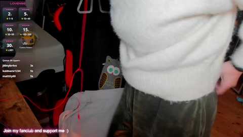 A video captures a person in a cozy room with a white sweater, green pants, and a red backpack, standing near a video game setup with a monitor and controller.