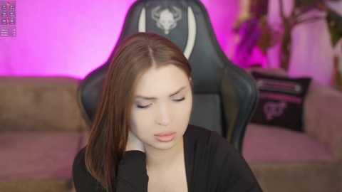 Video of a young woman with light skin and straight brown hair, wearing a black top, sitting in a black gaming chair with a skull emblem. Background features a pink-lit room with a beige sofa and a potted plant.