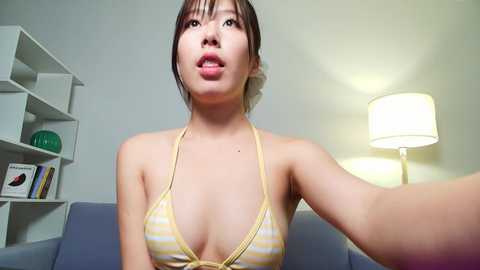 Video of an East Asian woman with light skin, brown hair, wearing a yellow and white striped bikini top, posing indoors with a lamp and bookshelf in the background.