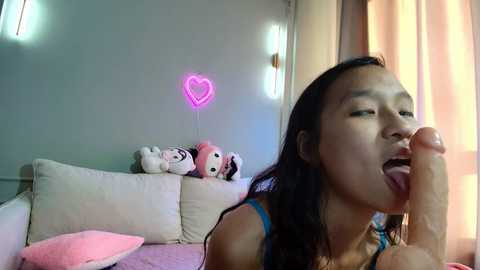 Video of an Asian woman with long black hair, wearing a blue top, licking a large erect penis. Background features a pink heart-shaped light, stuffed toys, and a beige couch.