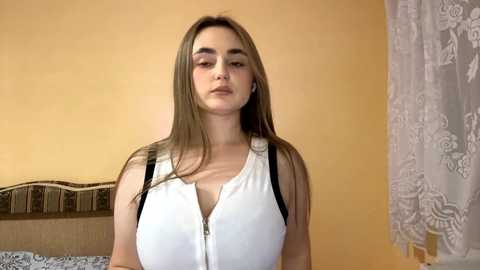 Video of a young, light-skinned woman with long, straight, light brown hair, wearing a white, sleeveless blouse with black straps, standing in a room with a beige wall and patterned curtains.