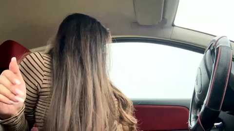 Video of a woman with long, straight, light brown hair, seated in a car, wearing a brown and beige striped long-sleeve shirt, giving a thumbs-up gesture, with the car's interior and window visible.