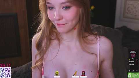 Video of a young, fair-skinned woman with long, wavy red hair, wearing a pink bra with small, cartoon figures on the cups. She has a slight smile and is indoors, surrounded by dim lighting.