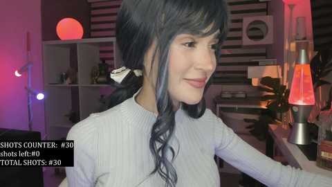 Video of a smiling woman with long, wavy dark hair, wearing a light gray sweater, in a dimly lit room with a red lamp, white shelves, and a green plant.