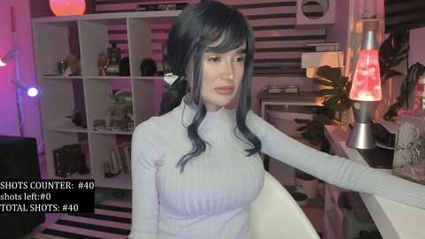 Video of a woman with long black hair in a light grey turtleneck sweater, sitting in a modern living room with white shelves, a red lamp, and a striped rug.