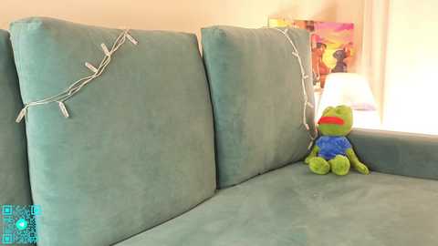 A video of a teal sofa with white wire-wrapped barbed wire design on the backrest, featuring a green frog toy wearing a blue shirt and red glasses, in a warmly lit room with a lamp and colorful artwork on the wall.