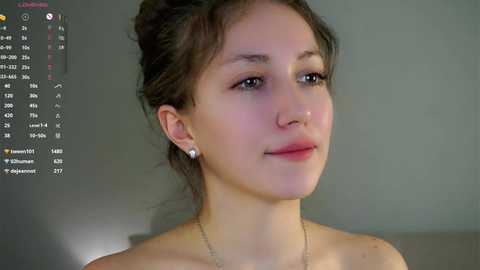 A close-up video of a young woman with fair skin, brown hair in a bun, wearing a delicate necklace, and small pearl earrings. She has a soft smile. Background shows a computer screen displaying a calendar.