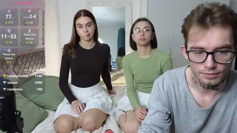 Media: Video of three young adults in a modern living room. Two women in casual attire, one in glasses, sit on a couch. A man in glasses stands in the foreground.