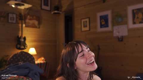 Video of a young woman with long brown hair, laughing joyfully in a cozy, dimly lit wooden-paneled room with framed art, guitar, and lamp.