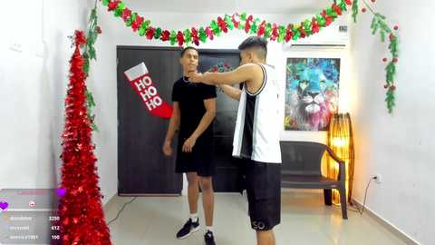 A video captures two young men, one in a black T-shirt, the other in a white and black sleeveless top, dancing in a festive room adorned with Christmas decorations and colorful posters.