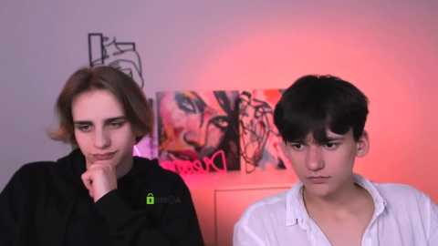 Video of two young people: a pale-skinned woman with shoulder-length brown hair, pensive expression, wearing a black jacket, and a darker-skinned man with short black hair, serious expression, in a white shirt. Background: abstract art and dim lighting.