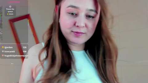 Video of a young, light-skinned woman with long, wavy brown hair, wearing a white tank top, standing against a beige brick wall, overlaid with social media filters and text.