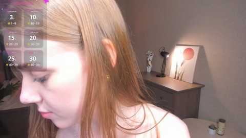 Video of a fair-skinned young woman with long, straight light brown hair, wearing a white top, in a dimly lit bedroom. Background includes a gray dresser, a lamp, and a framed abstract art piece.