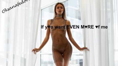 Video of a slender, light-skinned woman with long brown hair wearing a see-through fishnet bodysuit, standing in a bright, modern room with large windows and white curtains. Text overlay: \"If you want EVEN MORE <3 me.\