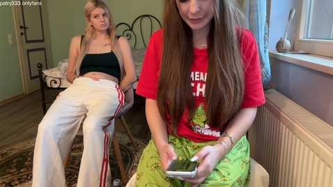 A video shows a blonde woman in a black tank top and white pants, sitting on a bed, while a brunette in a red shirt and green pants uses a phone near a window.