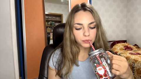 Video of a young woman with long brown hair, light skin, wearing a light blue shirt, drinking from a mason jar with a straw, in a cozy room with orange walls, bookshelves, and a patterned blanket.