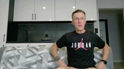 Video of a muscular, middle-aged man with short brown hair wearing a black t-shirt with white and red text and a black watch, sitting on a gray-patterned couch in a modern kitchen with white cabinets and a dark backsplash.