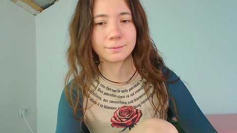 Video of a young woman with long brown hair, fair skin, wearing a beige shirt with a red rose graphic, in a room with a white wall and visible ceiling.