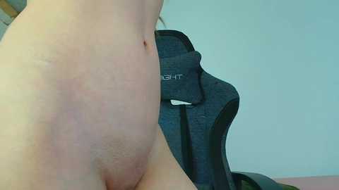 Video of a fair-skinned person's bare back, showing a red, slightly raised mark, seated on a grey gaming chair. The background is a plain, light blue wall.