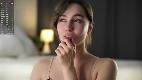 Media: A close-up video of a young woman with light skin, brown hair, and brown eyes, wearing a beige camisole, thoughtfully biting her finger in a dimly lit bedroom with a blurred lamp and bed.