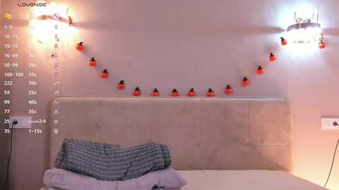 Video of a modern bedroom with a beige headboard, a blue and white striped blanket, and orange fairy lights on the wall.