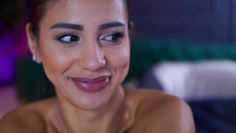 Video of a smiling, light-skinned woman with dark hair pulled back, wearing minimal makeup and a strapless top, against a blurred indoor background with green and purple hues.