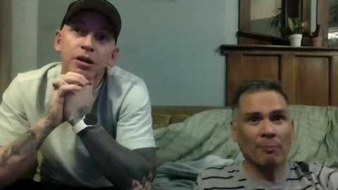 Video of two men watching TV; one wearing a baseball cap, grey shirt, and brace on his right arm, the other in a striped shirt. Background shows a wooden dresser and a potted plant.