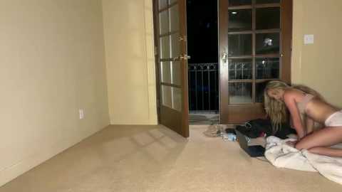 Media: Video of a blonde woman in lingerie kneeling on beige carpet in a dimly lit room with a partly open glass door, revealing a balcony outside.
